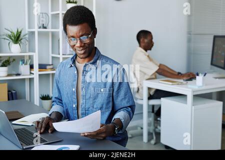 Portrait of young black man wearing glasses analyzing documents while working in office, copy space Stock Photo