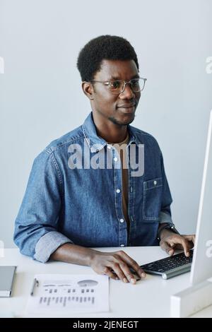 Vertical portrait of young black professional at workplace in office using PC computer against white wall Stock Photo