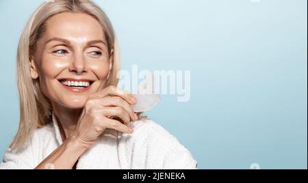Elegant smiling woman, 50 years old, using guasha tcm massaging tool, holding scrapper and looking aside at copy space, blue background Stock Photo