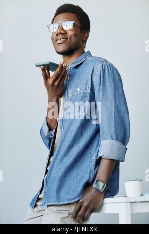 Vertical shot of young black man recording voice message via smartphone against minimal grey wall Stock Photo