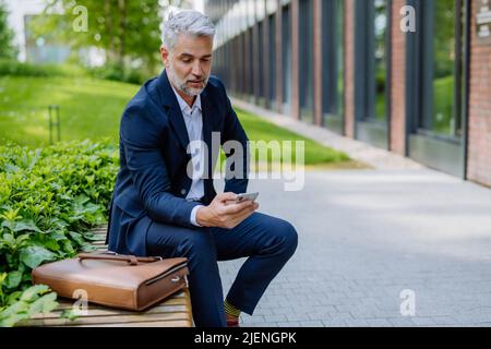 Successful mature businessman using smartphone while resting on street, sitting on bench and using mobile application. Stock Photo