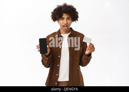 Portrait of shocked hispanic man shows credit card and mobile phone, screen app, standing in casual clothes over white background Stock Photo