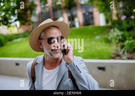 Confident man wearing straw hat and backpack talking on phone, businessman in casual clothes in summer on the way to work. Stock Photo