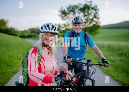 Active senior couple riding electric bicycles on road at summer park, healthy lifestyle concept. Stock Photo