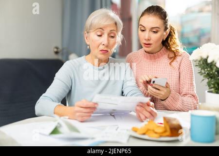 Elderly woman with adult daughter using smartphone and checking utility bills Stock Photo