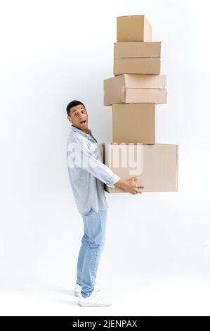 Excited mixed race guy in casual stylish clothes, delivery man, standing over isolated white background, holding many cardboard boxes, delivering parcels, looking in amazement at camera, delivery Stock Photo