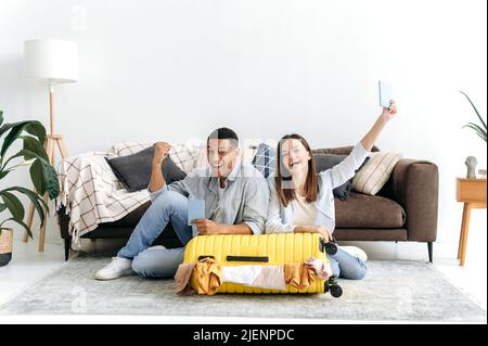 Cheerful multiracial couple, packed a trip suitcase for vacation, sit on the floor in living room near the yellow suitcase, hold their passports, look at camera, smile happily. Long-awaited vacation Stock Photo