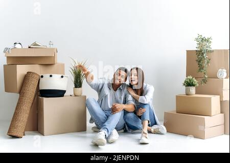 Happy young multiracial couple, caucasian woman and hispanic guy, are sitting on the floor between boxes of stuff in their new home or rental apartment, planning their house future design, smile Stock Photo