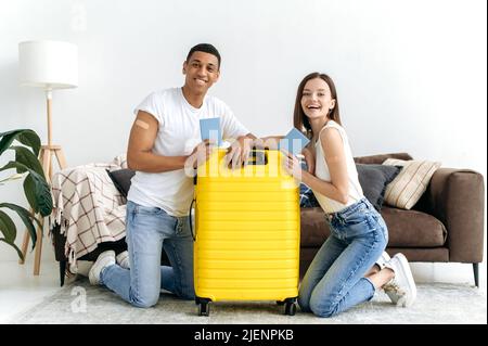 Long-awaited vacation, travel. Happy multiracial couple, got the vaccine and going on a trip, sit on the floor in the living room near the yellow suitcase, hold their passports, look at camera, smile Stock Photo