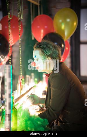 Clown concept - a man sitting in the dressing room with a paint and fake nose on his face and looking at his reflection in the mirror - holding a Stock Photo