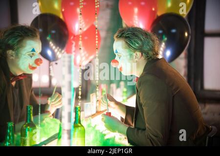 Clown concept - a man sitting in the dressing room with a paint and fake nose on his face and looking at his reflection in the mirror - holding a Stock Photo