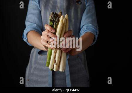 Crop unrecognizable female cook in apron holding bunch of raw healthy white and green asparagus against black background in studio Stock Photo