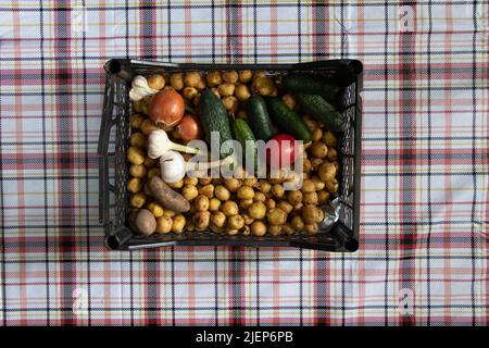 a box with vegetables, potatoes, onions, a tomato, standing on the table in the kitchen on a tablecloth in a cage, harvest from the garden, vegetables Stock Photo