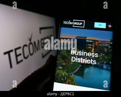 Person holding cellphone with webpage of Emirati real estate company Tecom Group LLC on screen in front of logo. Focus on center of phone display. Stock Photo