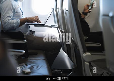 Businessman using laptop in airplane and passenger in front row with cellphone. Airline wifi connection, device flight mode concepts Stock Photo
