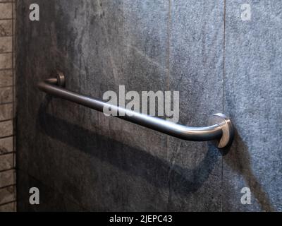 Close-up photo of stainless steel grab bar handrail installed on grey stone tiles wall background in a hotel handicapped disabled access bathroom. Stock Photo