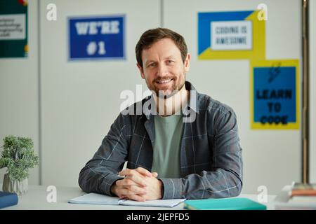 Front view portrait of smiling bearded teacher looking at camera while sitting at desk in school classroom against colorful posters Stock Photo