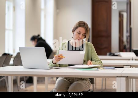 Hard-working female mature student reading architectural drawings while studying in public library Stock Photo