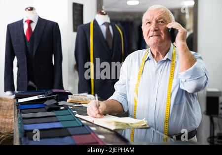 Professional tailor communicates with the client on mobile phone and takes notes in notebook Stock Photo