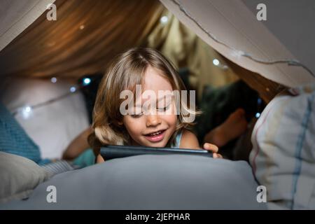 Happy cute little 6 year old boy having fun playing in teepee tent. Child using digital tablet watching cartoons or playing computer games lying in kid tent at home.  Stock Photo