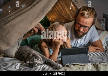 Portrait of a 6 year old boy and his father having fun playing in teepee tent. Father and son using digital tablet watching cartoons or playing computer games lying in kid tent at home.  Stock Photo
