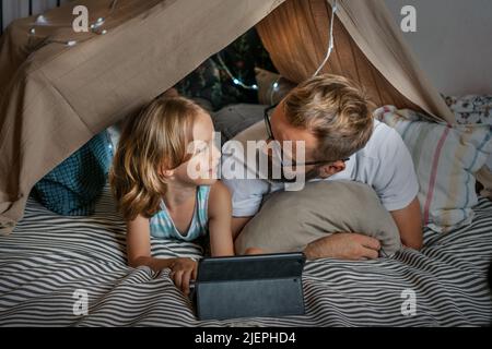 Portrait of a 6 year old boy and his father having fun playing in teepee tent. Father and son using digital tablet watching cartoons or playing computer games lying in kid tent at home.  Stock Photo