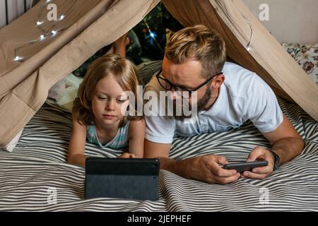 Portrait of a 6 year old boy and his father having fun playing in teepee tent. Father and son using digital tablet watching cartoons or playing computer games lying in kid tent at home.  Stock Photo