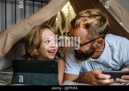 Portrait of a 6 year old boy and his father having fun playing in teepee tent. Father and son using digital tablet watching cartoons or playing computer games lying in kid tent at home.  Stock Photo
