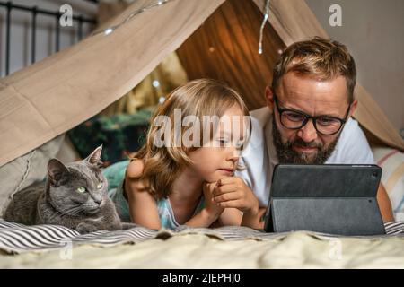 Portrait of a 6 year old boy and his father having fun playing in teepee tent. Father and son using digital tablet watching cartoons or playing computer games lying in kid tent at home.  Stock Photo