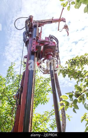 Photo of a drilling rig with a pipe for drinking water drilling against the background of trees and sky with clouds of warm sunny day. Stock Photo