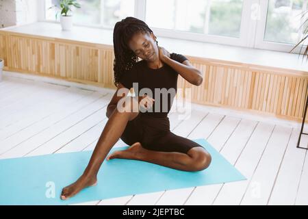Portrait dark skin multiracial woman with dreadlocks stretch, do yoga exercises on mat in gym. Rubbing body. Health care Stock Photo
