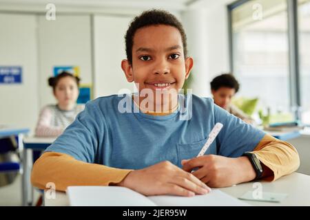 front view portrait of African American schoolboy looking at camera while sitting at desk in school classroom Stock Photo