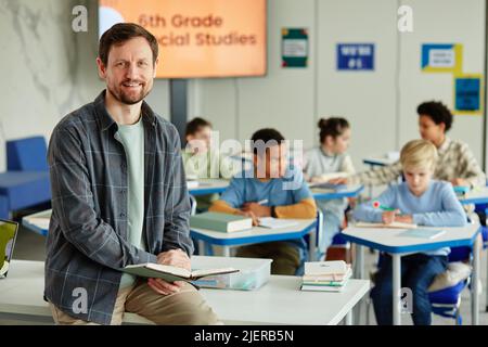 Portrait of bearded male teacher smiling at camera while standing in school classroom with children in background, copy space Stock Photo