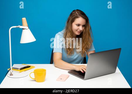 Young curly woman in a blue t-shirt studying from studio using laptop computer over a blue background. Serious beautiful young woman typing on laptop Stock Photo