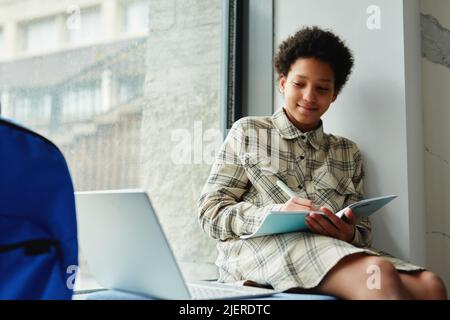 Portrait of smiling African American kid writing in notebook while sitting on window in school with laptop, copy space Stock Photo