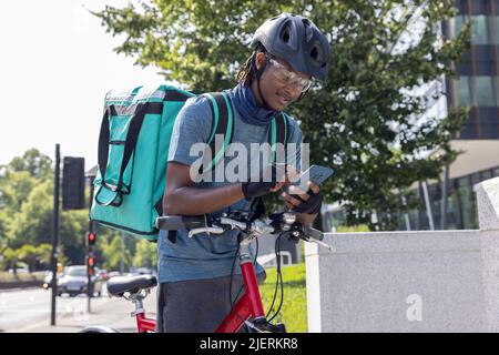 Courier On Bike Delivering Takeaway Food In City Checking Directions And Order Details On Mobile Phone Stock Photo