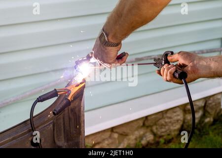 The hands of a man hold a welding machine and are engaged in welding metal and repairing a car door against the background of a country house. Men's t Stock Photo