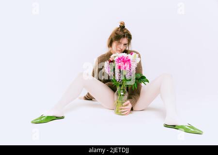 Young girl in oversized sweater and white tights posing with flowers isolated over grey studio background. Stock Photo