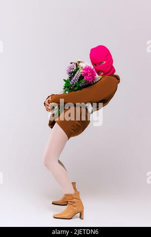 Portrait of stylish girl in oversized brown jacket, white tights, boots and pink neon balaclava posing with flowers isolated over grey background Stock Photo