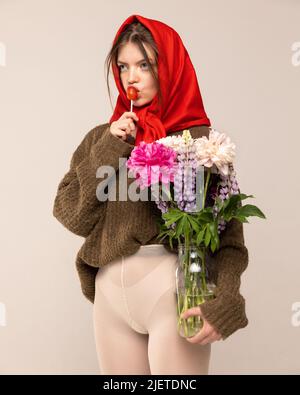 Portrait of young girl in red headscarf, sweater and white tights, posing with flowers and lollipop isolated over grey background Stock Photo