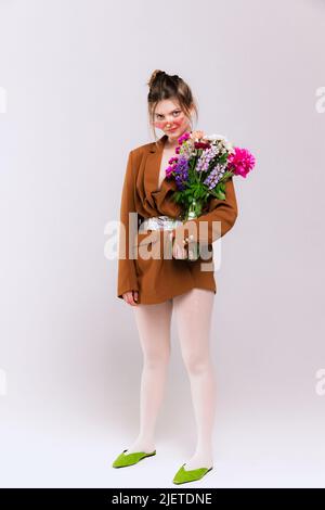 Portrait of stylish girl in oversized brown jacket, white tights and green retro shoes posing with flowers isolated over grey background Stock Photo
