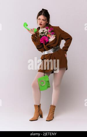 Portrait of stylish girl in brown jacket with flowers inside, white tights, boots posing with green vintage phone isolated over grey background Stock Photo