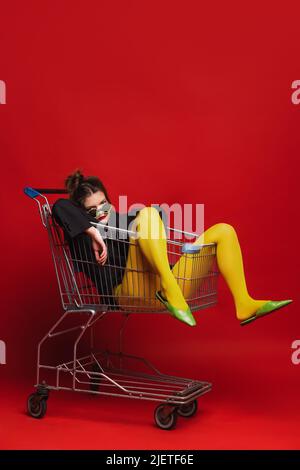 Funny portrait of stylish young girl in yellow tights sitting on shopping trolley, posing isolated over red background Stock Photo