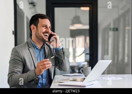 Cheerful male entrepreneur talking on smart phone. Young professional is holding eyeglasses while sitting at desk. He is wearing blazer while working Stock Photo
