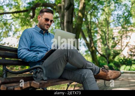 Confident young businessman in formals using laptop. Male professional is working on wireless technology. He is sitting on bench at public park. Stock Photo