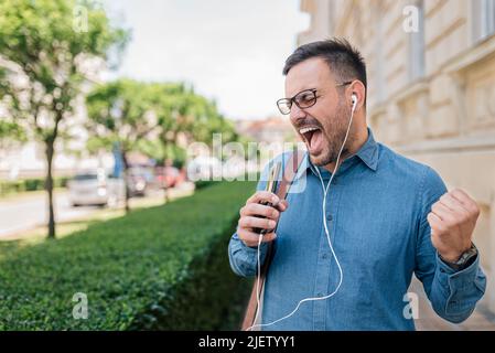 Entrepreneur wearing headphones screaming while clenching fist. Professional commuter is enjoying music through smart phone. He is walking on sidewalk Stock Photo