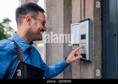 Smiling male executive using intercom by door. Young businessman is holding digital tablet. He is standing at office building entrance in the city. Stock Photo