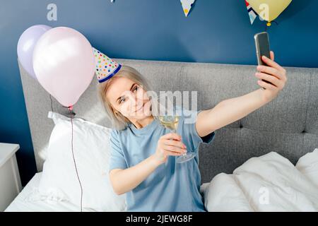 Happy emotional woman in pajama and party cap doing selfie shot on mobile phone at birthday morning while sitting on the bed with champagne and balloo Stock Photo