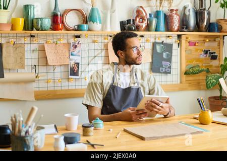 Young pensive black man in workwear using digital tablet while sitting by table in workshop and scrolling through online information Stock Photo