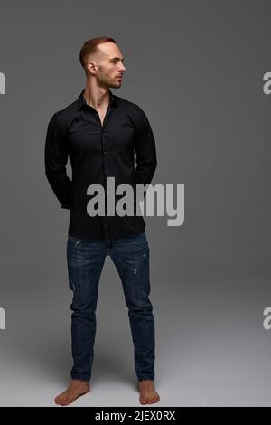 An attractive white man in jeans and black shirts tands in full growth against a gray background. Copy space Stock Photo